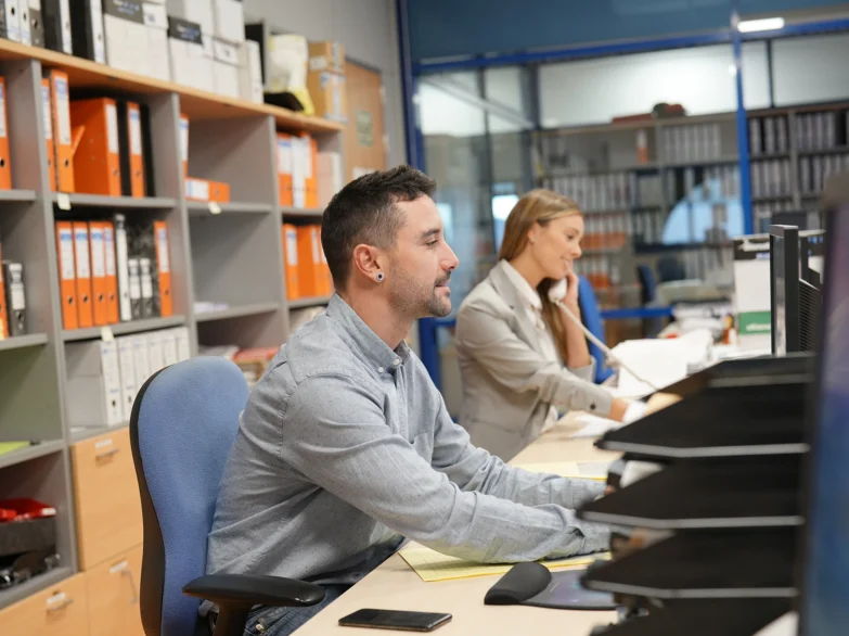 A man and woman collaborate at a desk in a modern office, surrounded by computers and paperwork.