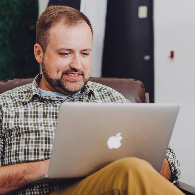 A man sitting in a chair, working on an Apple laptop, with a relaxed expression.