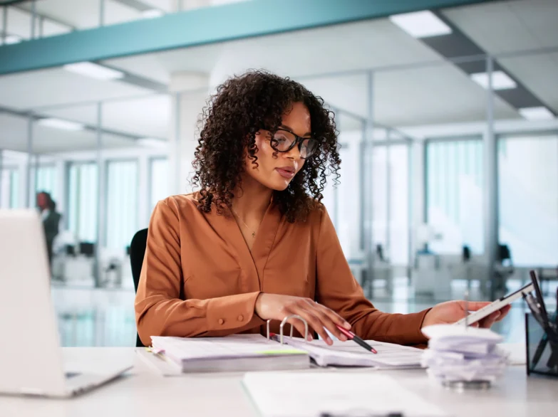 A woman wearing glasses sits at a desk, focused on her laptop, surrounded by books and a notepad.
