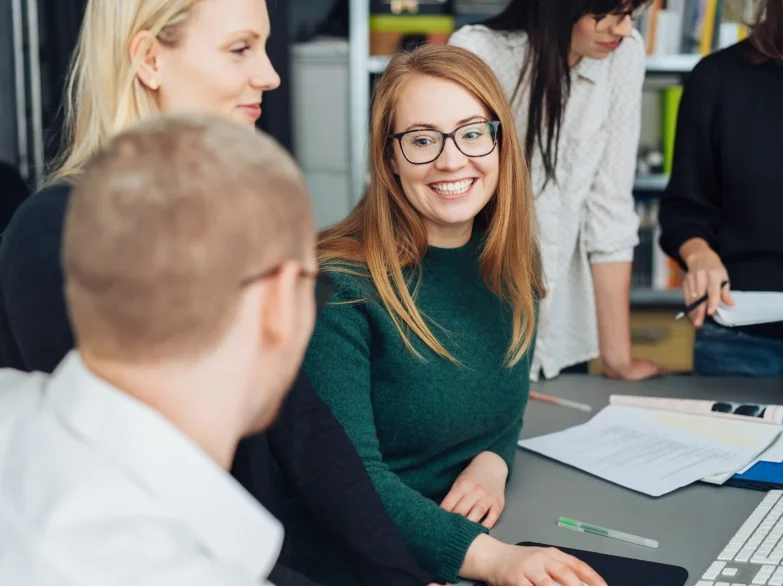 A group of people sitting around a table, with a smiling woman at the centre, enjoying a friendly conversation.