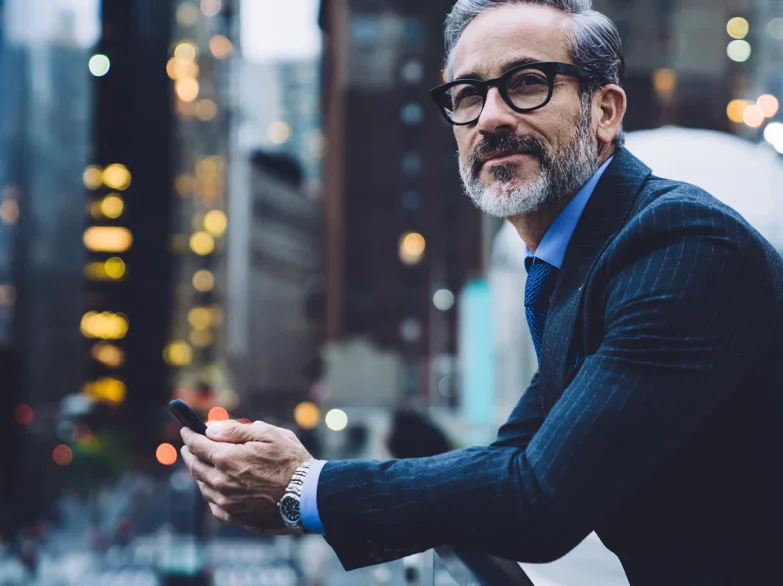 A man in a suit and glasses is focused on his phone with a city backdrop.