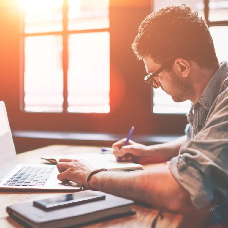 A man sits at a table, with a laptop, writing on a pad.
