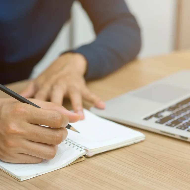 Man writing in a notebook with a pen, laptop open on a wooden table, focused on his work.
