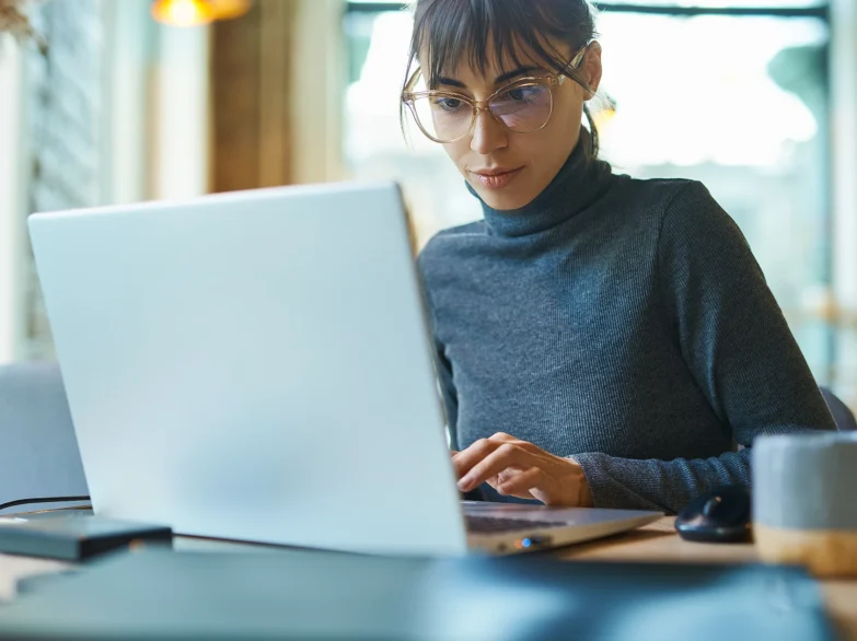 A woman wearing glasses is focused on her laptop, working intently in a well-lit environment.