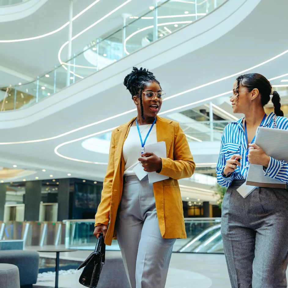 Two women walking together in a modern office building, engaged in conversation.
