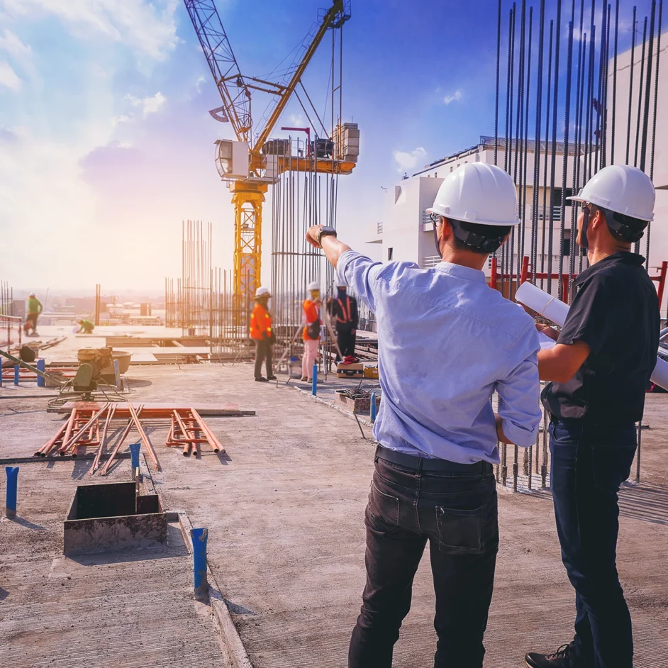 Two men in hard hats standing on a construction site, discussing plans with building materials in the background.