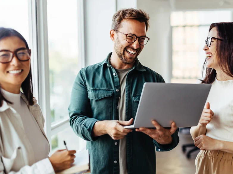 Three people in an office gather around a laptop, discussing something on the screen with happy expressions.
