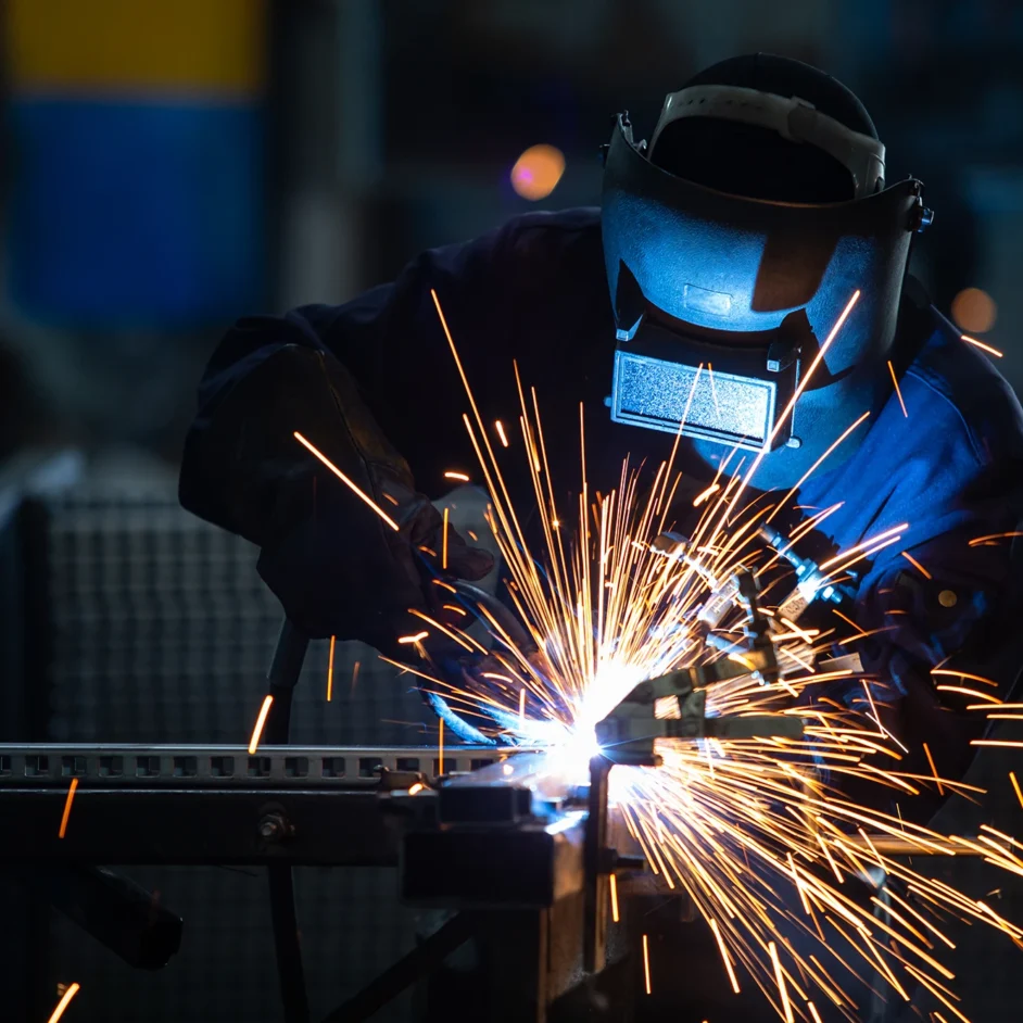 A man welding in a factory, surrounded by flying sparks and industrial equipment.