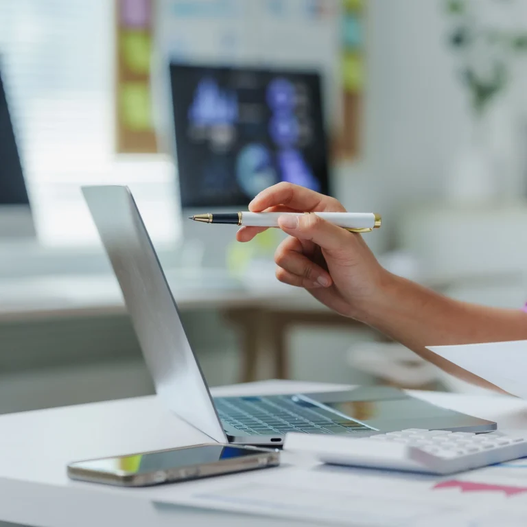 A woman sits at a desk with a pen in hand, focused on her laptop screen.