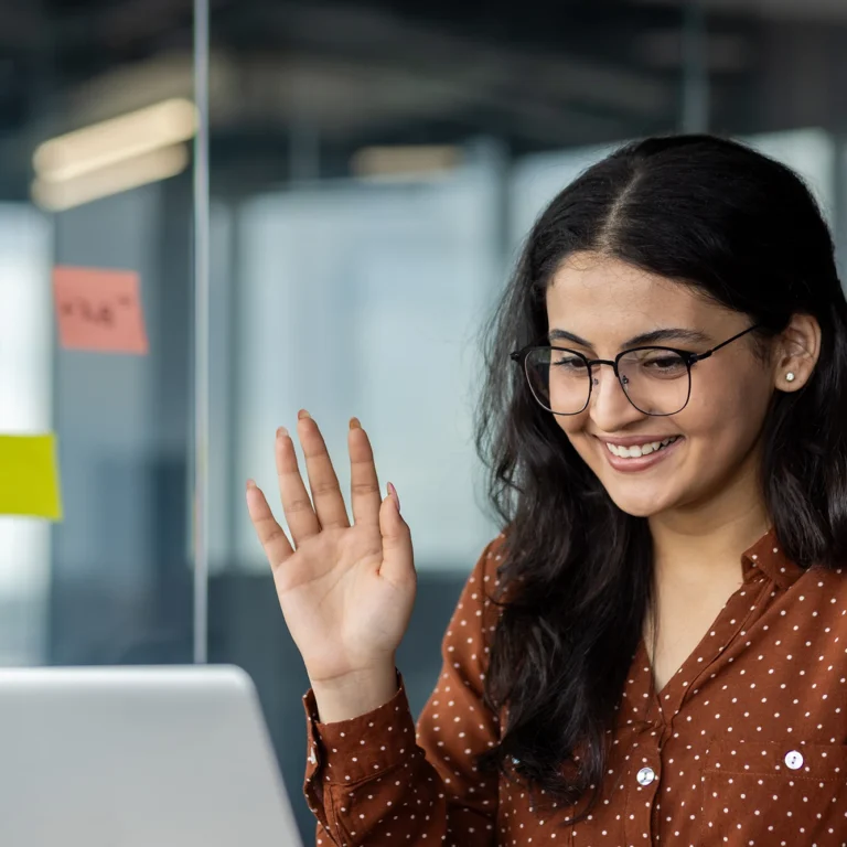 A woman waves cheerfully at her laptop screen, likely greeting someone during a video call.