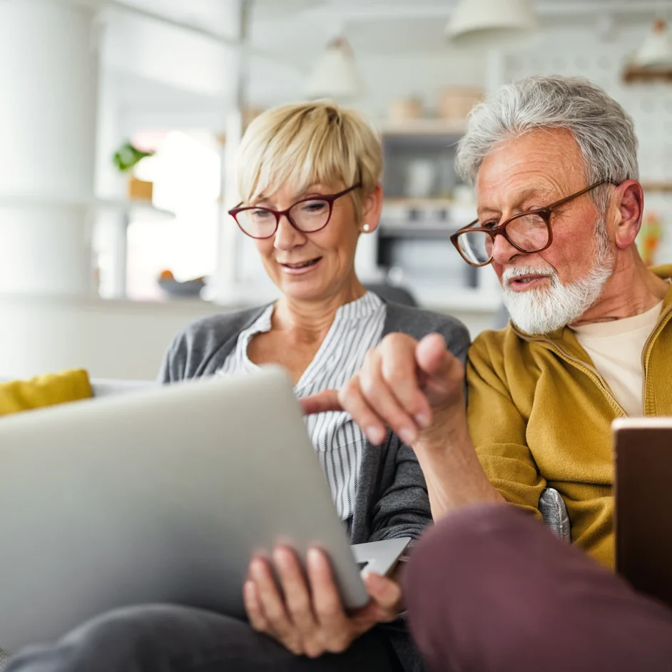 An older couple sitting together, smiling while using a laptop at home.