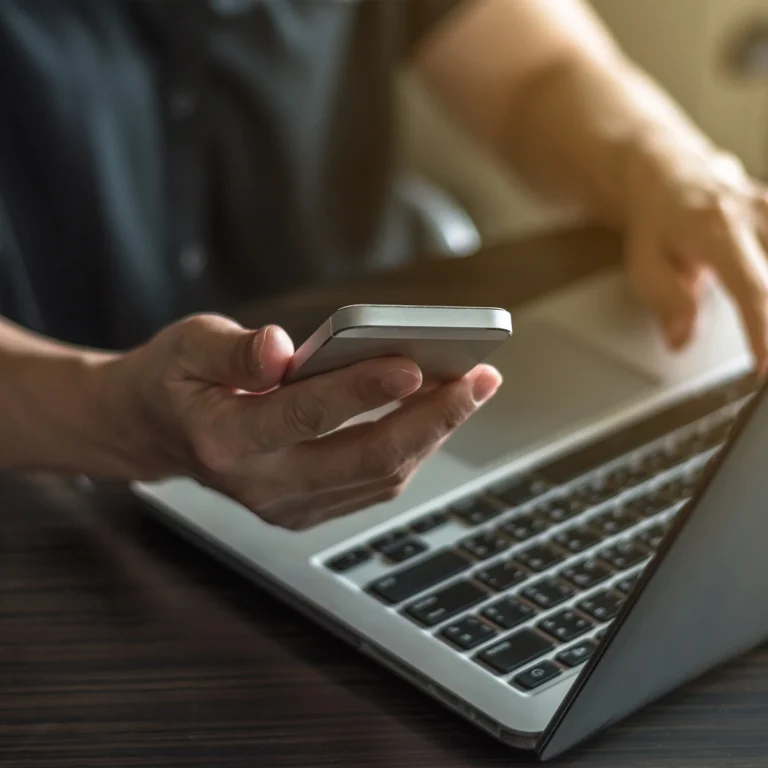 A person sitting at a desk, using a cell phone in one hand and typing on a laptop with the other.