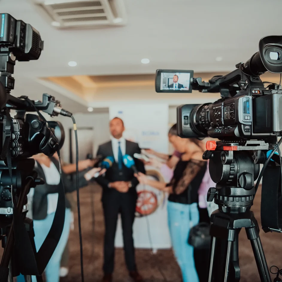 A man in a suit stands confidently in front of a camera, ready to speak.