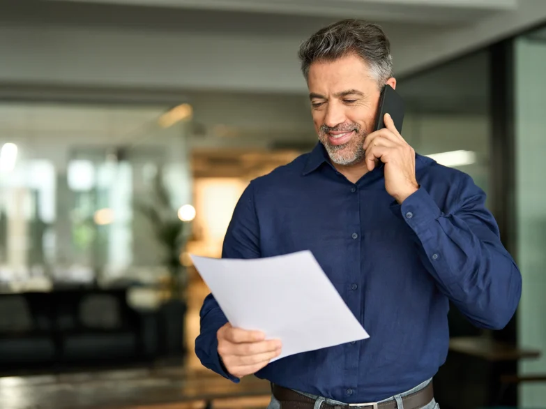 A man talks on the phone while holding a piece of paper in his other hand, appearing engaged in a conversation.