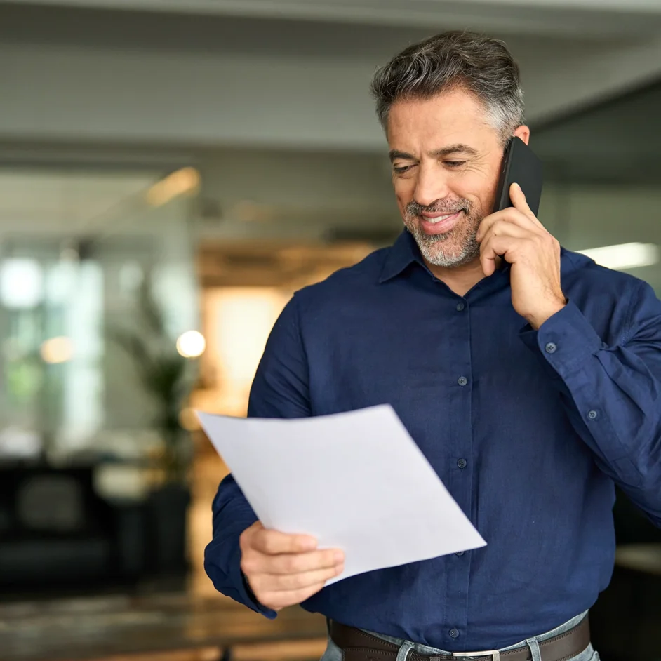 A man talks on the phone while holding a piece of paper in his other hand, appearing engaged in a conversation.