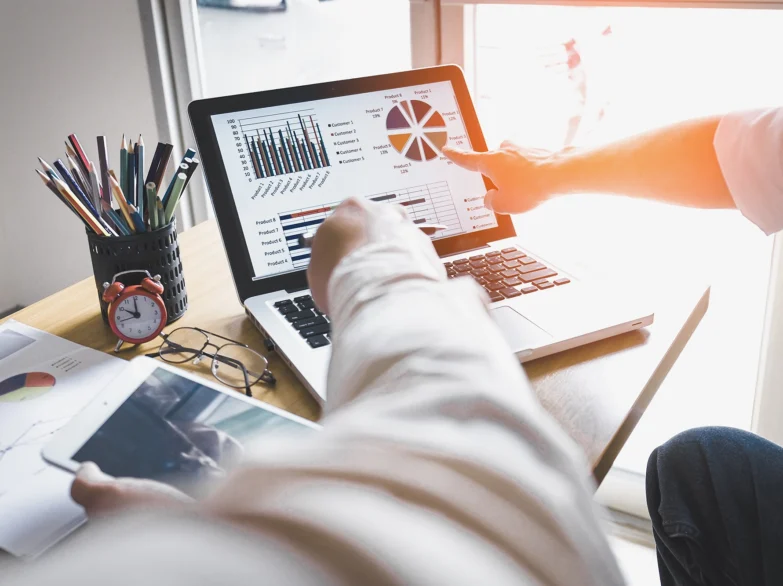 Two people collaborating at a desk with a laptop and scattered papers, engaged in discussion and work.