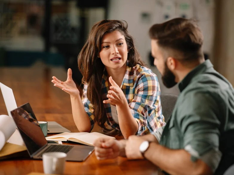 A man and woman sit at a table, each using a laptop, engaged in conversation and collaboration.