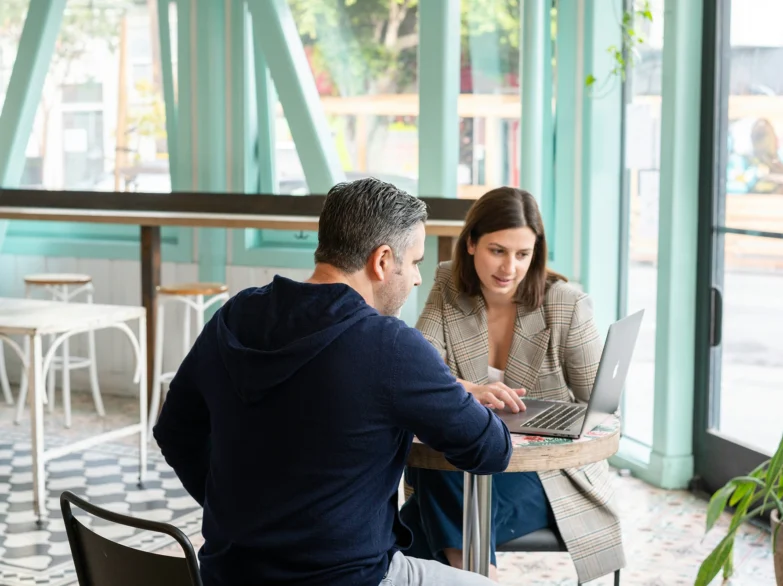 A man and woman sit at a table, focused on a laptop, engaged in conversation or collaboration.