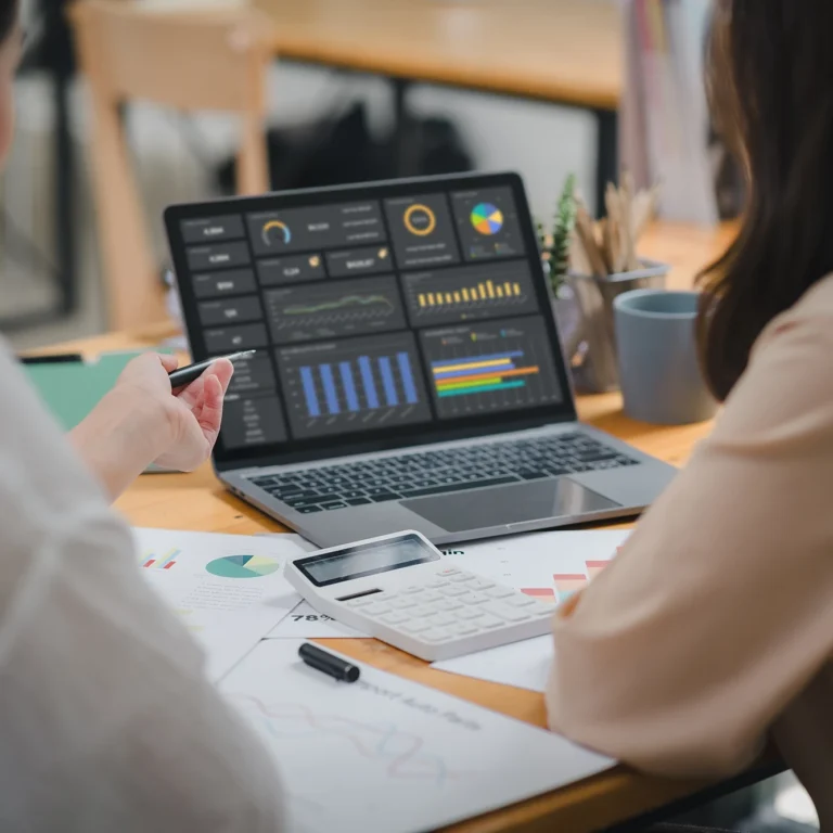 Two women collaborate at a table, working on a laptop surrounded by papers.