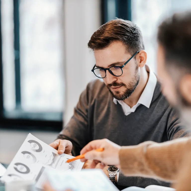 Two men collaborate on a project, discussing ideas and working together at a table with laptops and papers.
