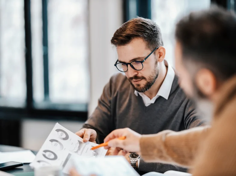 Two men collaborate on a project, discussing ideas and working together at a table with laptops and papers.