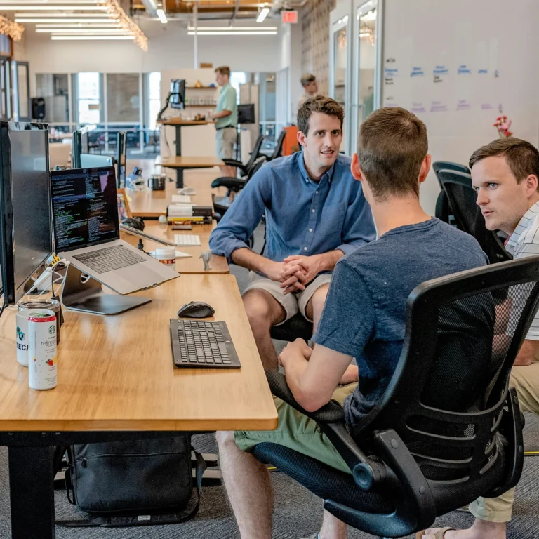 Three men engaged in conversation while sitting at a table in a modern office setting.