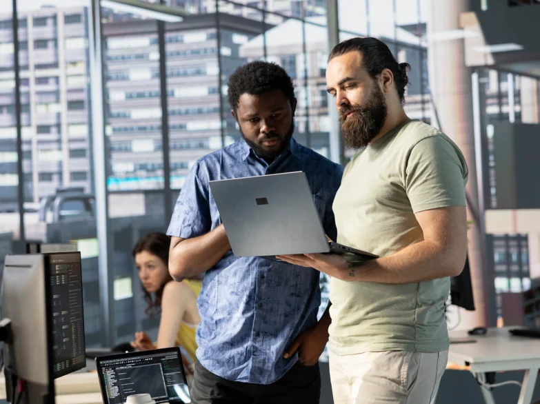 Two men standing together, looking at a laptop computer on a table, engaged in conversation.